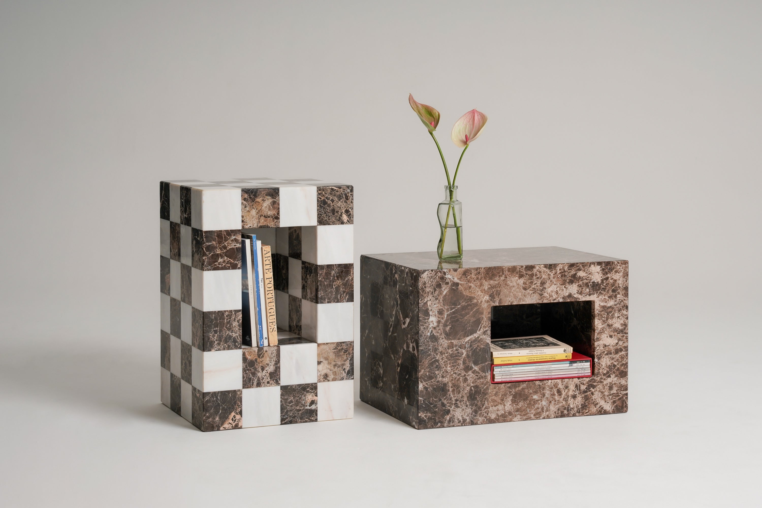Two modern rectangular side tables or coffee tables on a light gray studio background. One table has a black-and-white checkerboard pattern and is called Pipoca, and the other is made of brown marble and is called Trufa with a recessed shelf holding books.