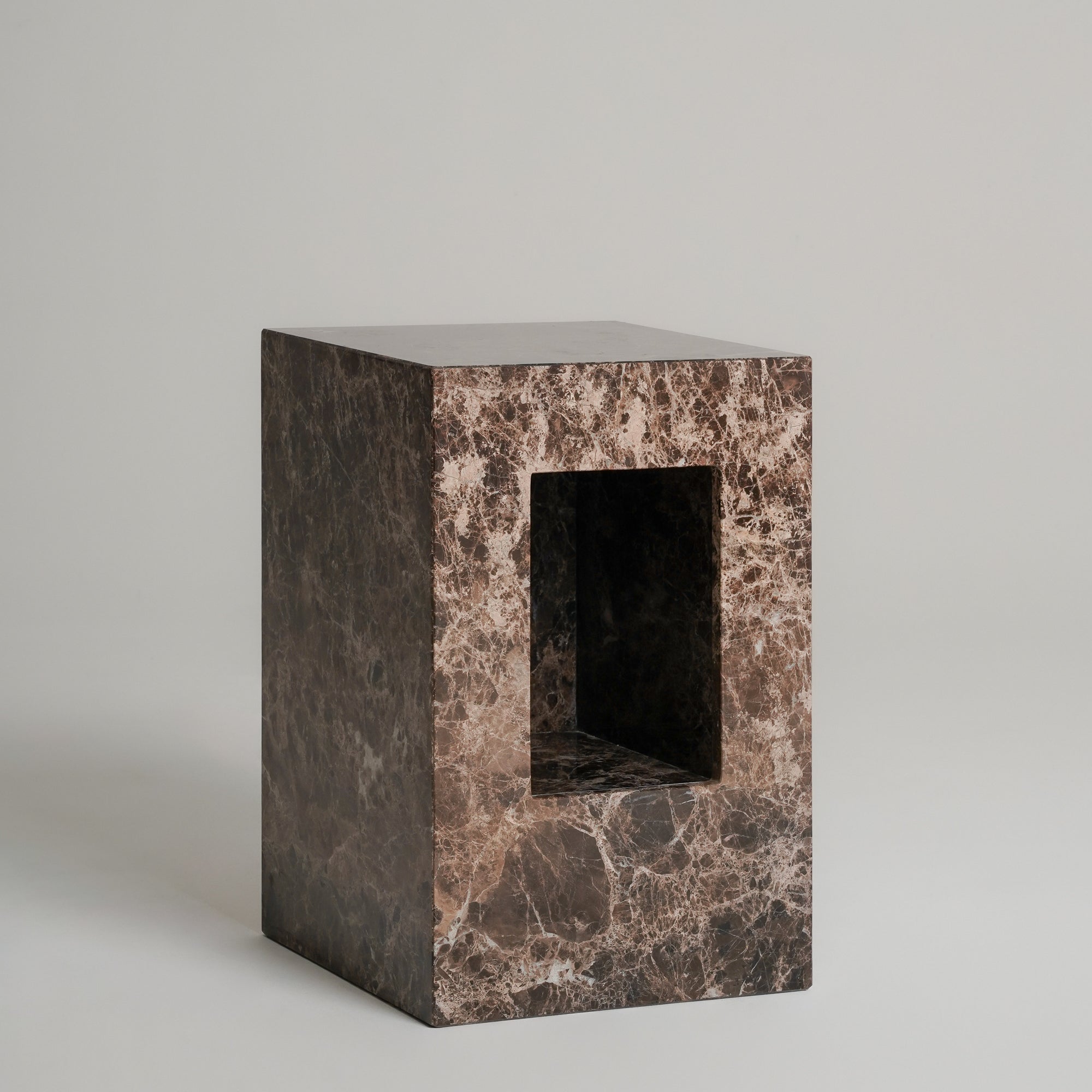 A rectangular stone pedestal or block with a central rectangular cutout, made from dark brown and beige marbled stone, photographed on a neutral light gray background.