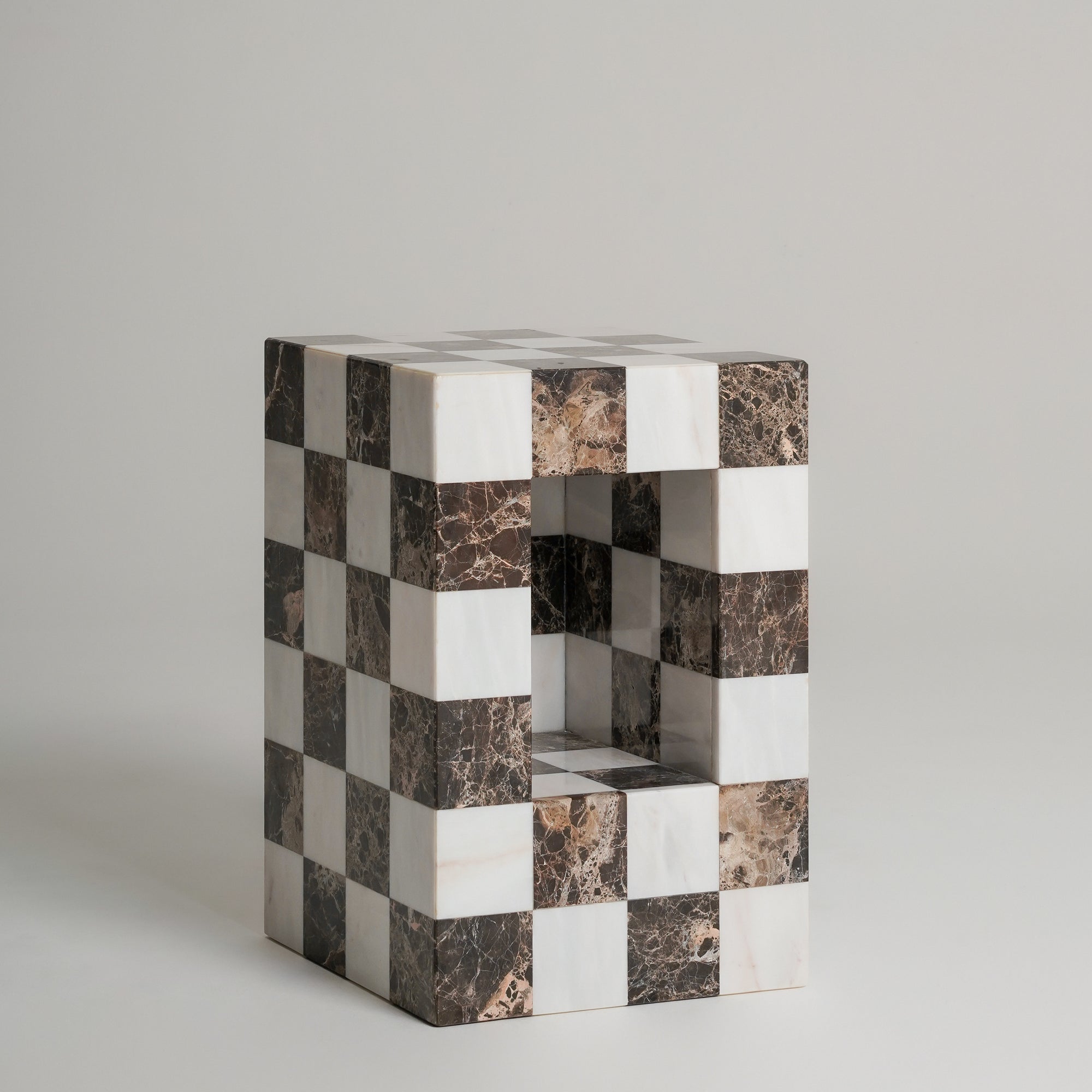 A small rectangular decorative object with a checkerboard pattern of white and dark brown marble tiles, featuring an open center, set against a plain light gray background.