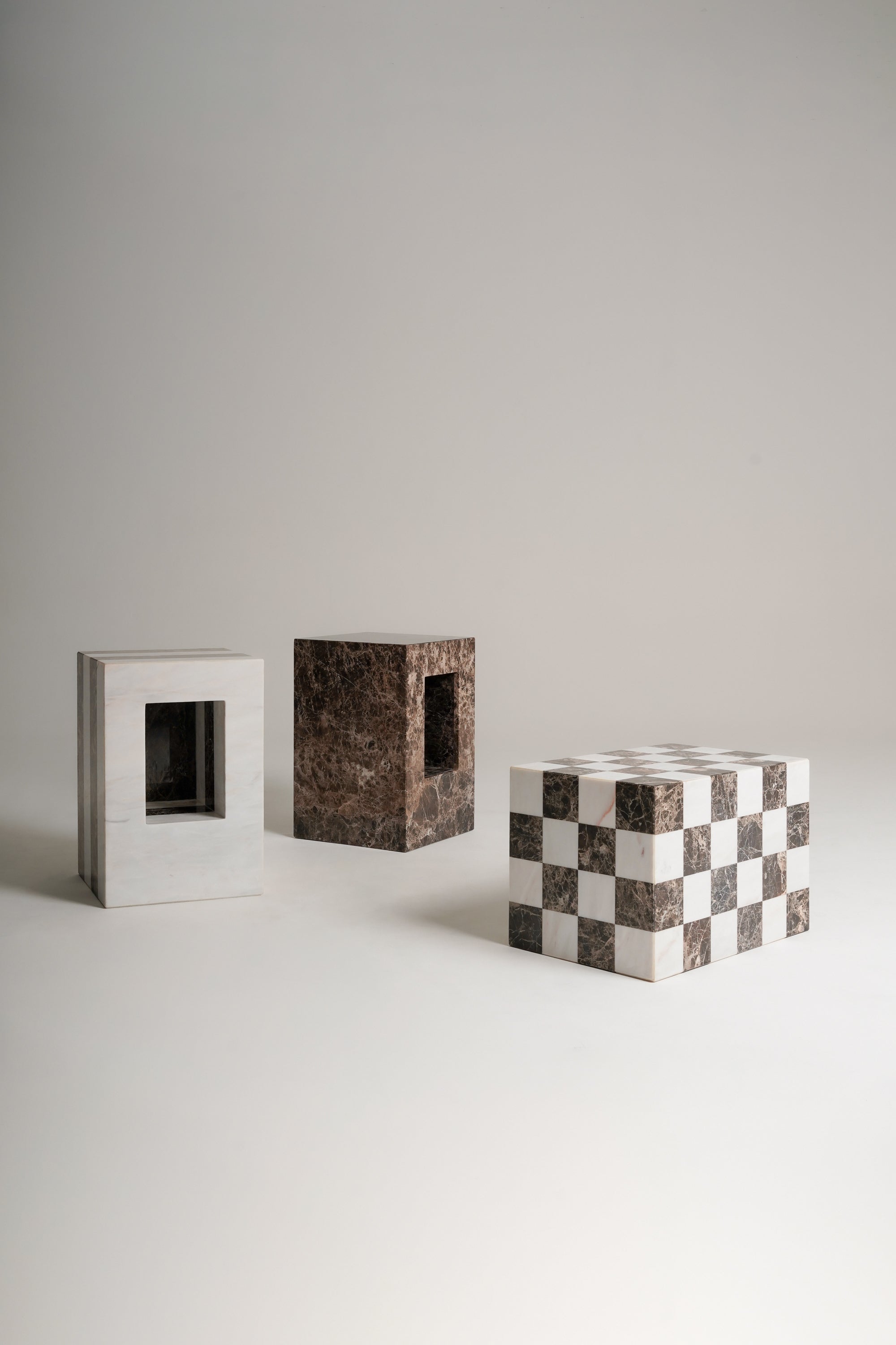 Three rectangular sculptures made of marble stone are arranged on a light gray studio floor against a matching gray background. One sculpture is striped with brown and white marble, one is dark brown marbled, and one is a white and dark brown checkerboard pattern.
