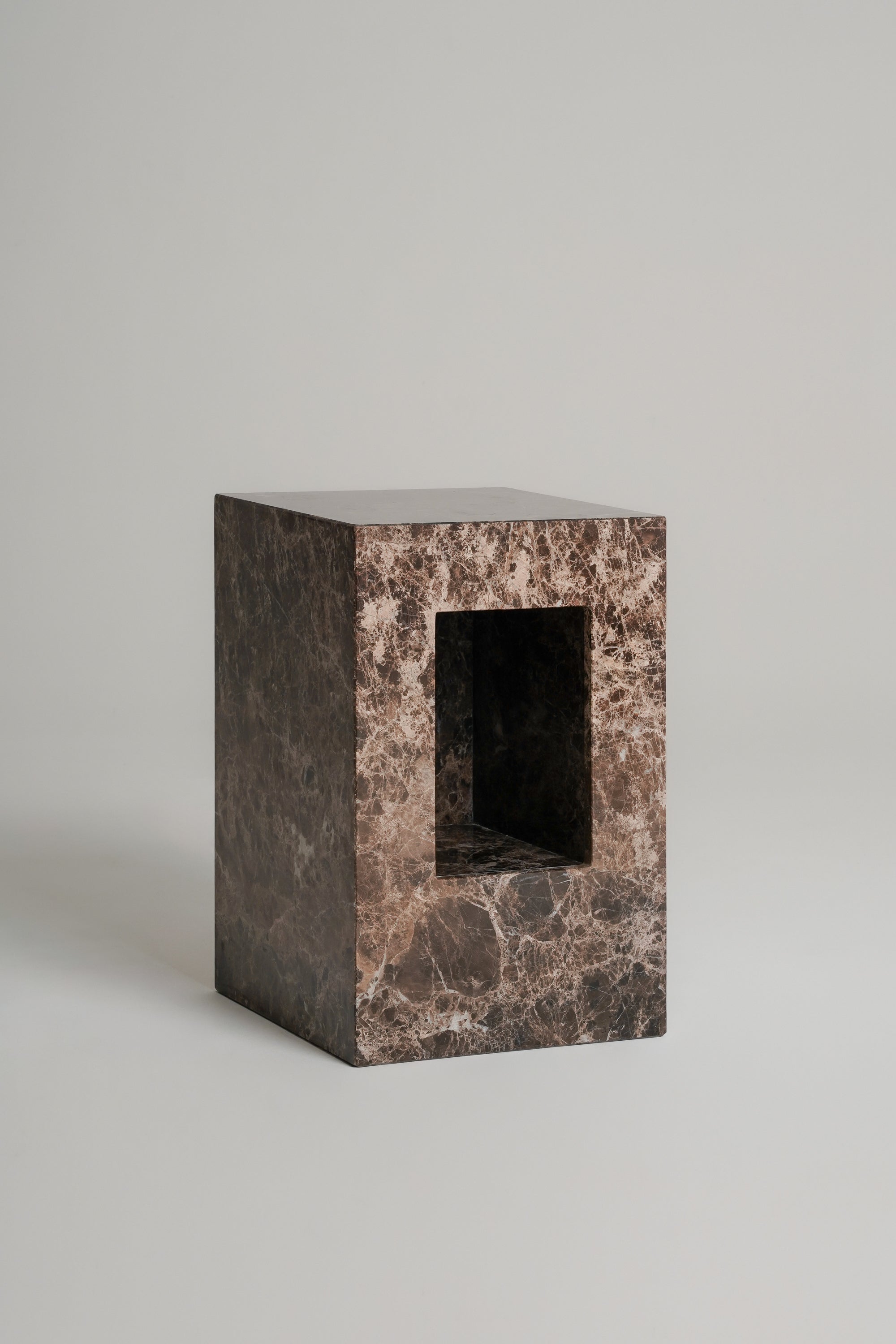 A rectangular stone pedestal or block with a central rectangular cutout, made from dark brown and beige marbled stone, photographed on a neutral light gray background.