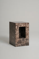 A rectangular stone pedestal or block with a central rectangular cutout, made from dark brown and beige marbled stone, photographed on a neutral light gray background.