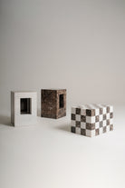 Three rectangular sculptures made of marble stone are arranged on a light gray studio floor against a matching gray background. One sculpture is striped with brown and white marble, one is dark brown marbled, and one is a white and dark brown checkerboard pattern.